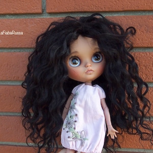 May include: A doll with large eyes and long, black, curly hair. The doll is wearing a light pink dress with floral detailing. The doll's face has delicate features. The doll is positioned in front of a brick wall.