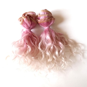 Peut inclure: Deux perruques de poupée avec des cheveux roses et blancs ombrés. Les perruques sont coiffées avec un chignon sur le dessus et de longs cheveux ondulés en cascade. Les chignons ont une bordure dorée.