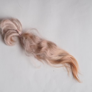 Peut inclure: Un paquet de fibres de laine teintes en rose clair et marron, nouées à une extrémité. Les fibres sont longues et ondulées, avec un dégradé de couleurs allant du rose clair au marron plus foncé aux extrémités. La laine est douce et duveteuse.