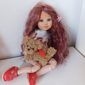 Könnte beinhalten: Eine Puppe mit langem, welligem malvenfarbenem Haar, blauen Augen und hellem Teint, bekleidet mit einem ärmellosen weißen Oberteil mit rotem Blumenmuster und roten Sandalen. Die Puppe hält einen kleinen, flauschigen beigen Teddybären mit roter Schleife.