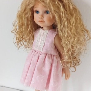 Peut inclure: Une poupée aux longs cheveux blonds bouclés et volumineux avec des yeux bleu vif. La poupée porte une robe rose clair sans manches ornée d'un motif circulaire blanc répétitif et d'une bordure en dentelle blanche verticale sur le corsage.