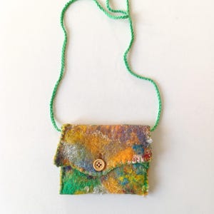 Peut inclure: Un petit sac bandoulière en feutre coloré avec une fermeture à bouton et une sangle en cordon vert. Le sac présente un mélange de teintes jaunes, oranges, vertes et bleues, créant un aspect texturé et artistique. Le sac mesure environ 13 cm de large.
