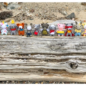 May include: A row of 12 knitted animal figurines, each wearing a different outfit. The figurines are arranged on a piece of driftwood. The animals include a unicorn, a fox, a pig, a bee, a giraffe, and a mouse.