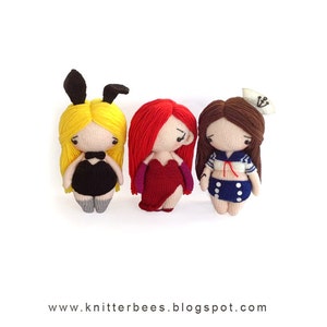 May include: Three crocheted dolls dressed in different costumes. The doll on the left is wearing a black bunny costume with a yellow wig. The doll in the middle is wearing a red dress with a red wig. The doll on the right is wearing a blue and white sailor outfit with a brown wig.