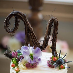 May include: A two-tiered wedding cake decorated with white frosting and a delicate, wavy pattern. The cake is topped with a twig letter 'M' and a variety of purple, white, and red flowers.