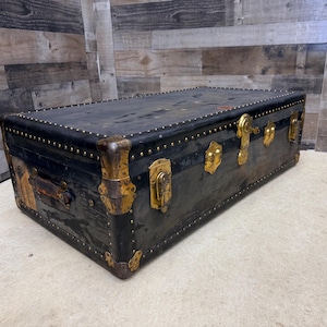 May include: A vintage black trunk with brass hardware and studded accents. The trunk features a rectangular shape with a hinged lid, corner protectors, and a handle. The trunk's dimensions are approximately 36 inches long, 20 inches wide, and 20 inches high.