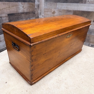 Op de afbeelding: Een houten opbergkist met een gebogen deksel en een warme, amberkleurige afwerking. De kist heeft een metalen handvat aan de zijkant en een decoratieve sluiting aan de voorkant. De houtnerf is zichtbaar.