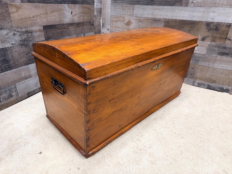 Op de afbeelding: Een houten opbergkist met een gebogen deksel en een warme, amberkleurige afwerking. De kist heeft een metalen handvat aan de zijkant en een decoratieve sluiting aan de voorkant. De houtnerf is zichtbaar.