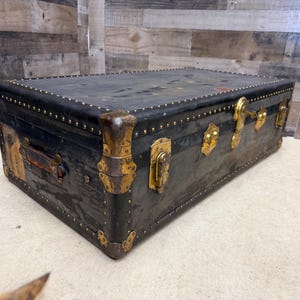 May include: A vintage black trunk with brass hardware, including corner protectors, latches, and studs. The trunk has a leather handle and shows signs of wear, suggesting its age and use. The trunk is rectangular and sits on a light-colored surface.
