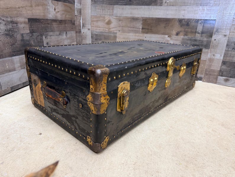 May include: A vintage black trunk with brass hardware, including corner protectors, latches, and studs. The trunk has a leather handle and shows signs of wear, suggesting its age and use. The trunk is rectangular and sits on a light-colored surface.