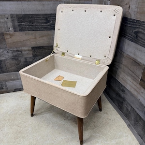 May include: A vintage sewing box with a hinged lid, upholstered in a light beige fabric. The box sits on four wooden legs and features brass hinges. The interior is also beige, with a small tag inside. A classic storage solution.