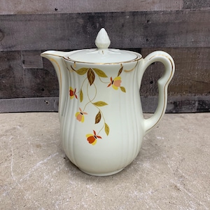 May include: A white ceramic teapot with a gold trim and a floral design featuring brown, orange, and yellow leaves and flowers. The teapot has a lid with a knob on top.