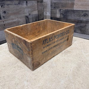 May include: A weathered wooden crate with the text "ATLAS POWDER CO." printed in black. The crate is rectangular with a visible dovetail joint construction. The background is a light-colored surface and a wooden wall.