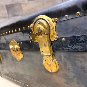 May include: Close-up of an antique trunk with a black exterior and brass hardware. The trunk features a key lock and decorative rivets. The brass hardware shows signs of age and wear, adding to its vintage appeal.