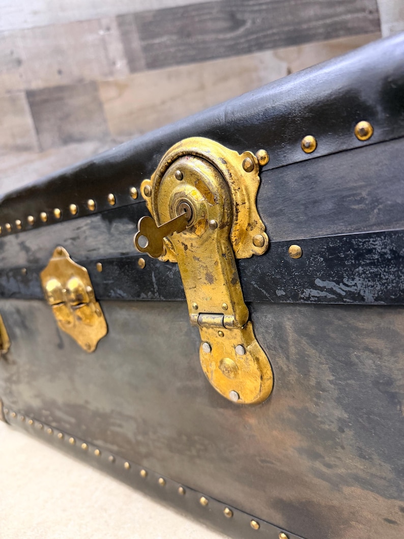 May include: Close-up of an antique trunk with a black exterior and brass hardware. The trunk features a key lock and decorative rivets. The brass hardware shows signs of age and wear, adding to its vintage appeal.
