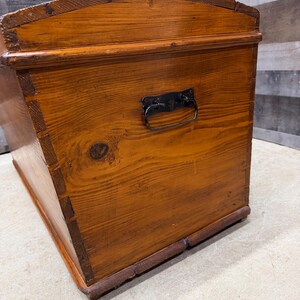 Op de afbeelding: Een vintage houten kist met een warme, roodbruine afwerking. De kist heeft een gebogen bovenkant, een metalen handvat en zichtbare houtnerf. De hoeken zijn verstevigd met donkerder hout, wat wijst op een klassieke opbergoplossing.