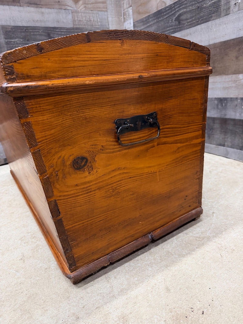Op de afbeelding: Een vintage houten kist met een warme, roodbruine afwerking. De kist heeft een gebogen bovenkant, een metalen handvat en zichtbare houtnerf. De hoeken zijn verstevigd met donkerder hout, wat wijst op een klassieke opbergoplossing.