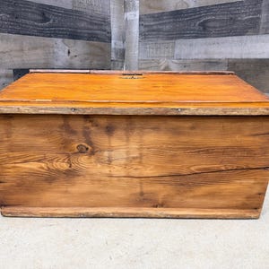 Op de afbeelding: Een rechthoekige houten opbergkist met een scharnierend deksel. De kist is gemaakt van houten planken en heeft een warme, oranjebruine afwerking. De houtnerf is zichtbaar en de kist lijkt verouderd.