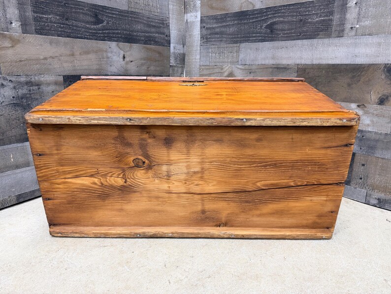 Op de afbeelding: Een rechthoekige houten opbergkist met een scharnierend deksel. De kist is gemaakt van houten planken en heeft een warme, oranjebruine afwerking. De houtnerf is zichtbaar en de kist lijkt verouderd.