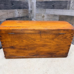 Op de afbeelding: Een rechthoekige houten kist met een gebogen deksel. De kist is gemaakt van lichtbruin hout met zichtbare nerfpatronen en metalen scharnieren. De kist wordt waarschijnlijk gebruikt voor opslag.