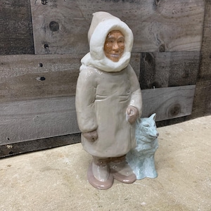 May include: A ceramic figurine of a person wearing a white hooded parka and holding a white wolf. The figurine is standing on a light brown surface.