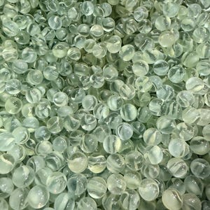 May include: A close-up shot of a pile of clear glass marbles with a slight green tint. Each marble has a swirled, opaque white pattern inside. The marbles are smooth and round, filling the frame.