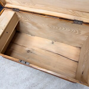 Op de afbeelding: Open houten kist met een lichtbruine afwerking. De binnenkant is gemaakt van lichtgekleurd hout met zichtbare nerf en een klein compartiment aan de linkerkant. De kist heeft metalen scharnieren en grendels.