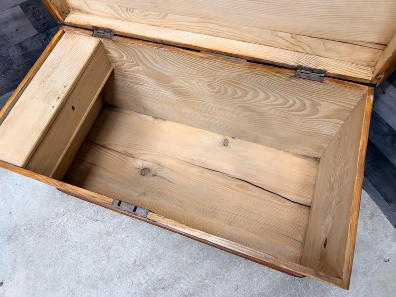 Op de afbeelding: Open houten kist met een lichtbruine afwerking. De binnenkant is gemaakt van lichtgekleurd hout met zichtbare nerf en een klein compartiment aan de linkerkant. De kist heeft metalen scharnieren en grendels.
