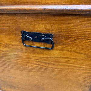 Op de afbeelding: Close-up van een houten kist met een donkerbruine afwerking. De kist heeft een zwarte metalen handgreep en hardware. De houtnerf is zichtbaar en de kist lijkt vintage of antiek.