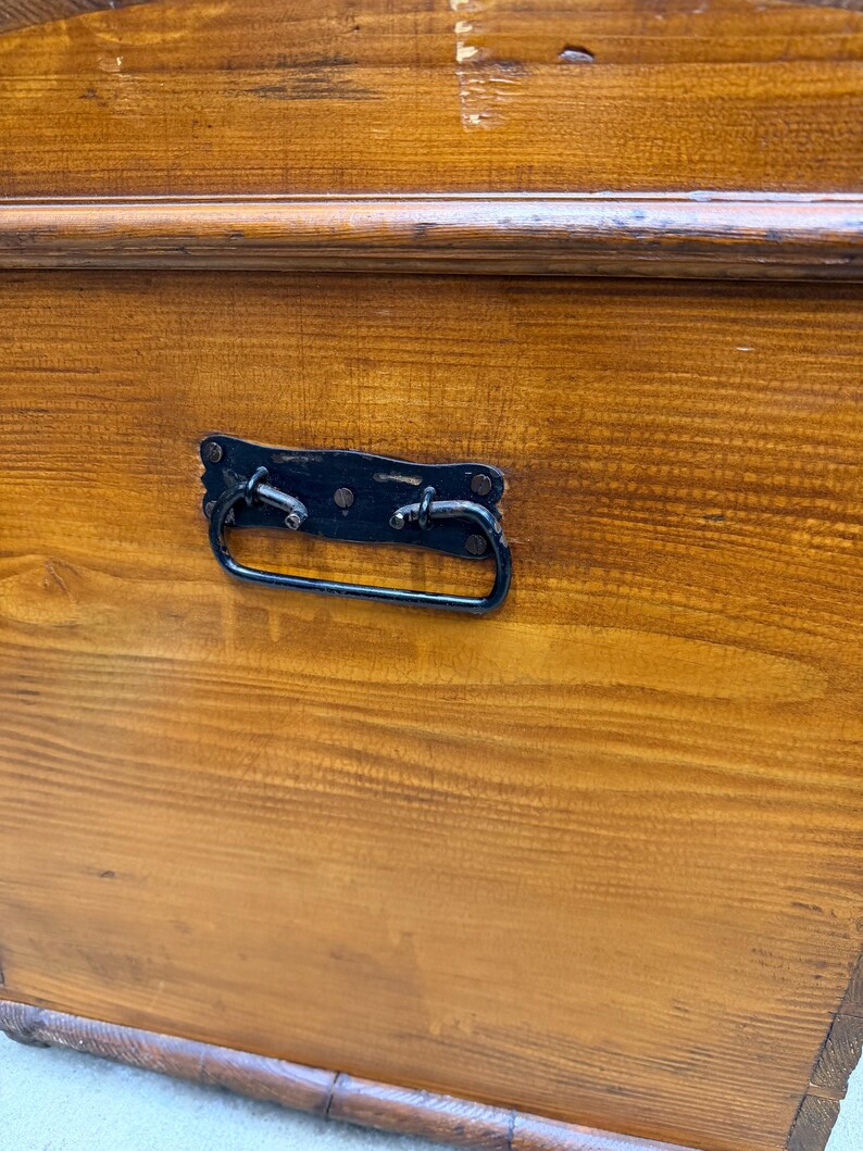 Op de afbeelding: Close-up van een houten kist met een donkerbruine afwerking. De kist heeft een zwarte metalen handgreep en hardware. De houtnerf is zichtbaar en de kist lijkt vintage of antiek.