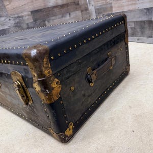 May include: An antique black trunk with brass accents. The trunk features a leather handle, brass corner protectors, and numerous brass studs. The trunk's exterior shows signs of wear, adding to its vintage appeal. The trunk is rectangular in shape.