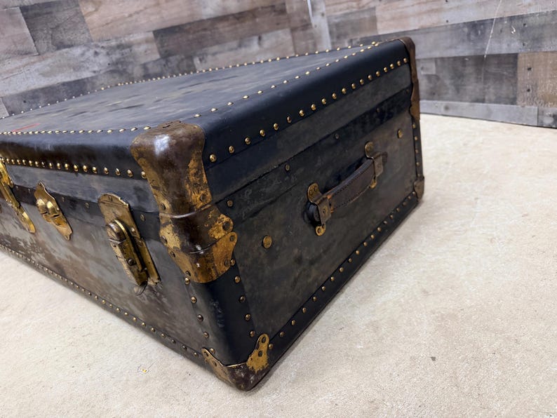 May include: An antique black trunk with brass accents. The trunk features a leather handle, brass corner protectors, and numerous brass studs. The trunk's exterior shows signs of wear, adding to its vintage appeal. The trunk is rectangular in shape.