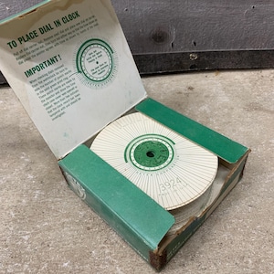 May include: An open cardboard box with a green and white clock dial inside. The box has green trim and text that reads "TO PLACE DIAL IN CLOCK" and "IMPORTANT!". The dial has the number "3924" printed on it.