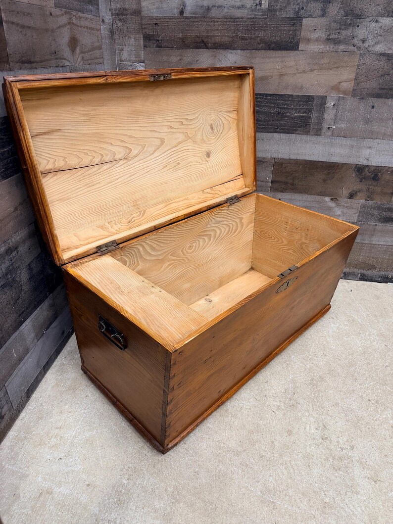 Op de afbeelding: Open houten kist met een scharnierend deksel. De kist is gemaakt van lichtbruin hout met zichtbare nerfpatronen. Metalen hardware, inclusief scharnieren en een handvat, is zichtbaar. De binnenkant is ook lichtbruin.
