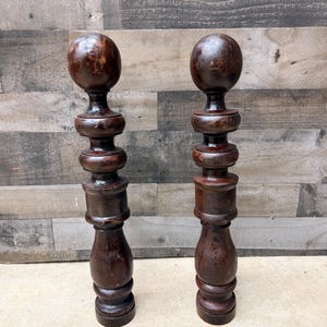 May include: Two dark brown wooden balusters featuring a turned design with a spherical top finial, a series of stacked circular rings, a central cylindrical section, and a flared base. The wood has a polished finish with visible grain patterns.