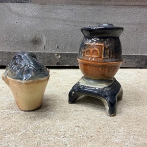 May include: Two ceramic pieces: a small, dark blue and tan salt shaker and a miniature stove-shaped container. The stove has a black top, a brown midsection, and sits on a black base. The salt shaker is a light tan with a dark blue top.