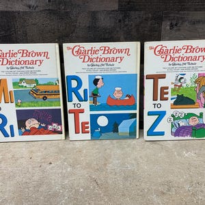 May include: Three vintage hardcover books titled "The Charlie Brown Dictionary" with illustrations from the Peanuts comic strip. The books are divided into three sections: M to R, R to Te, and Te to Z.