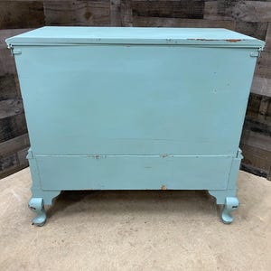 Pu&ograve; includere: Uno scrigno in legno blu chiaro con una finitura usurata. Lo scrigno ha un coperchio incernierato e poggia su quattro gambe tornite.