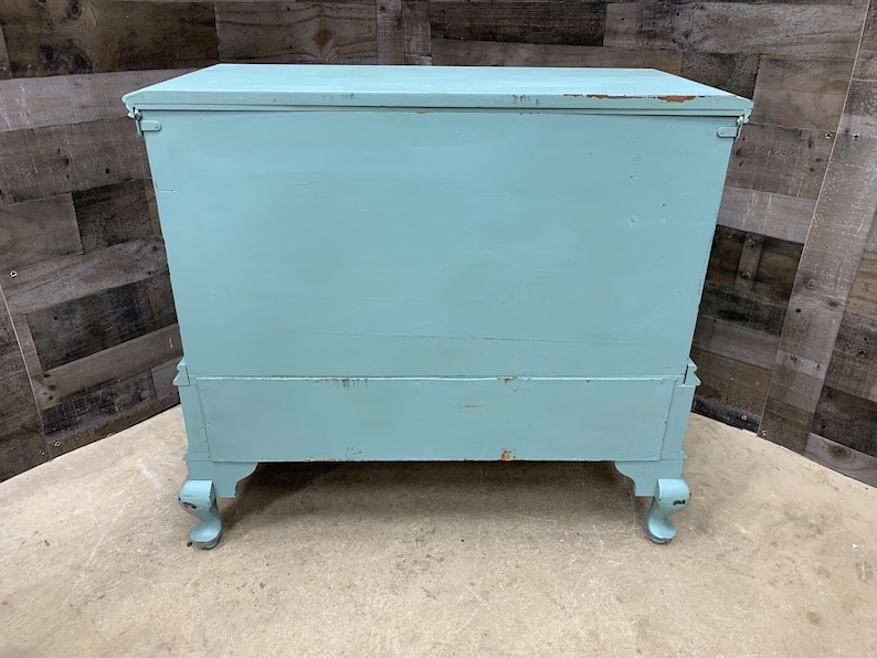 Pu&ograve; includere: Uno scrigno in legno blu chiaro con una finitura usurata. Lo scrigno ha un coperchio incernierato e poggia su quattro gambe tornite.
