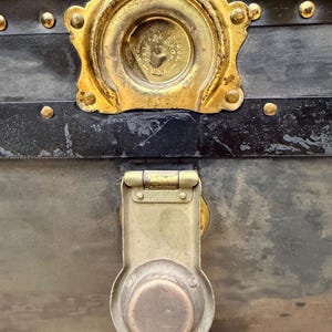 May include: Close-up of an antique trunk with a black exterior and brass hardware. The trunk features a large, ornate lock and a decorative brass plate. Rivets and metal accents add to the vintage aesthetic.