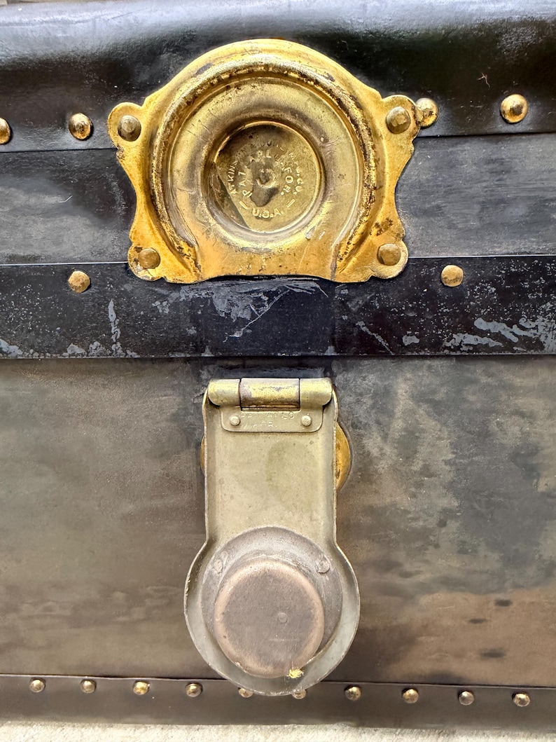 May include: Close-up of an antique trunk with a black exterior and brass hardware. The trunk features a large, ornate lock and a decorative brass plate. Rivets and metal accents add to the vintage aesthetic.