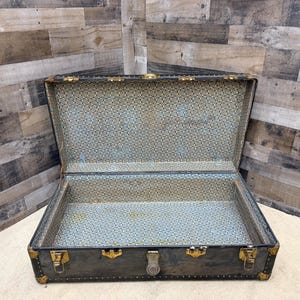 May include: An open, antique trunk with a dark exterior and gold-colored hardware. The interior is lined with a patterned fabric in shades of blue, tan, and gold. The trunk is rectangular with a hinged lid and metal clasps.