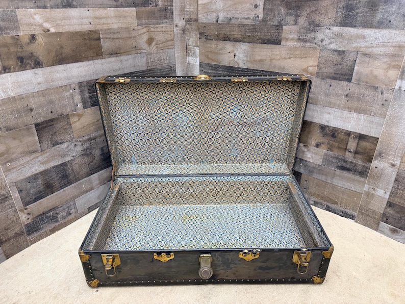 May include: An open, antique trunk with a dark exterior and gold-colored hardware. The interior is lined with a patterned fabric in shades of blue, tan, and gold. The trunk is rectangular with a hinged lid and metal clasps.
