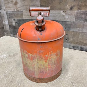 May include: An orange vintage metal gas can with a handle and a spout. The can has a cylindrical shape with a rounded top and a rusty base. The can shows signs of wear and tear, with some areas of paint loss and discoloration.