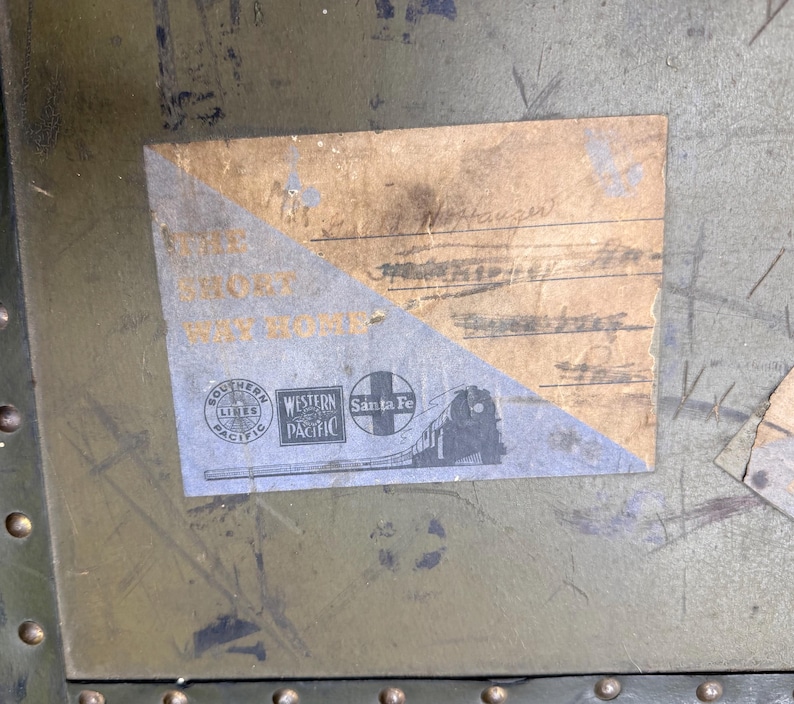 Może przedstawiać: Stara etykieta bagażowa z tekstem i logo kolei Southern Pacific, Western Pacific i Santa Fe. Etykieta ma postarzany, wyblakły wygląd i jest przymocowana do zielonej metalowej powierzchni.