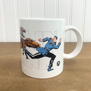 May include: White ceramic mug with a graphic of a man running from a dinosaur. The text "DINO RUN" is printed on the mug.