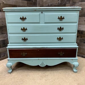 Pu&ograve; includere: Un com&ograve; di colore azzurro chiaro con cinque cassetti e ferramenta in ottone. Il com&ograve; ha una finitura invecchiata e poggia su quattro gambe a cavaliere.