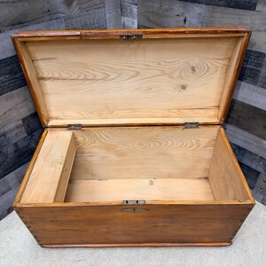 Op de afbeelding: Open houten kist met een scharnierend deksel. De kist is gemaakt van lichtgekleurd hout met een warme, bruine vlek. Het interieur toont de natuurlijke houtnerf. Metalen scharnieren en een grendel beveiligen het deksel.