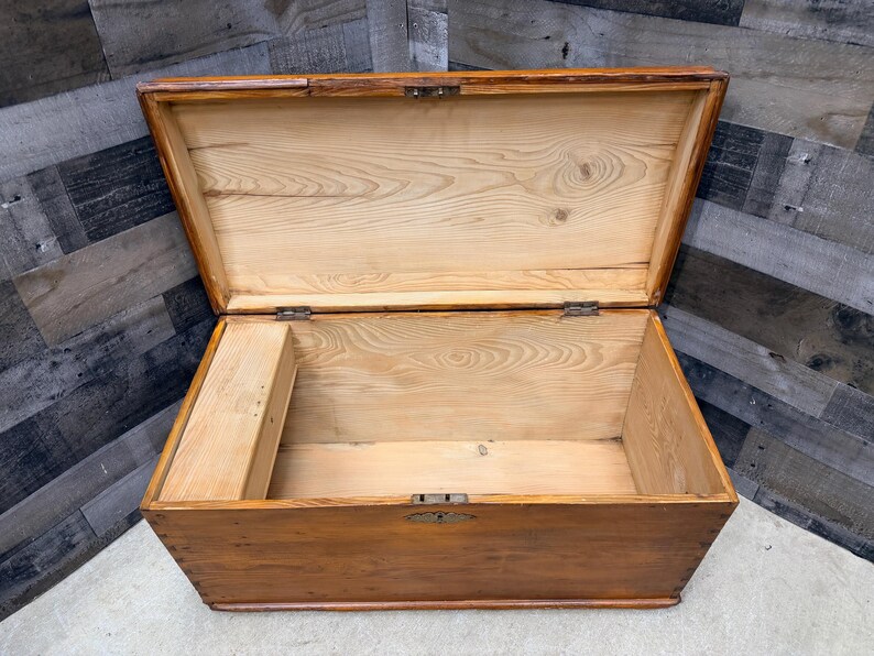 Op de afbeelding: Open houten kist met een scharnierend deksel. De kist is gemaakt van lichtgekleurd hout met een warme, bruine vlek. Het interieur toont de natuurlijke houtnerf. Metalen scharnieren en een grendel beveiligen het deksel.