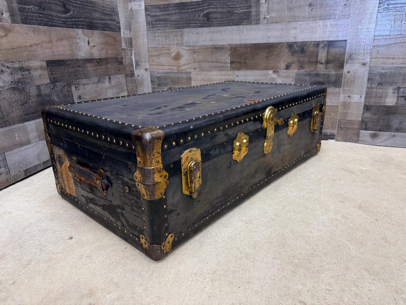 May include: A vintage black trunk with brass hardware and studded accents. The trunk features a rectangular shape with a hinged lid, corner protectors, and a handle. The trunk's dimensions are approximately 36 inches long, 20 inches wide, and 20 inches high.